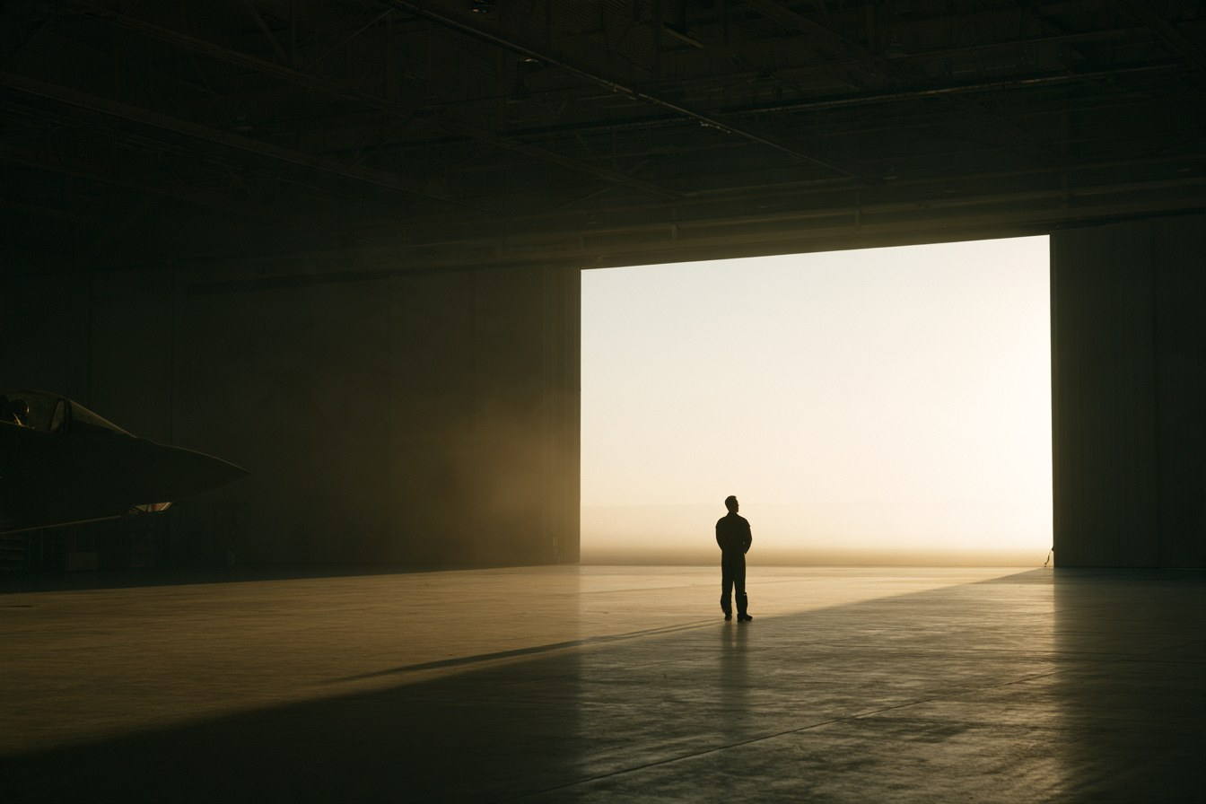 A pilot standing alone in a vast hangar, facing the open door and the light.